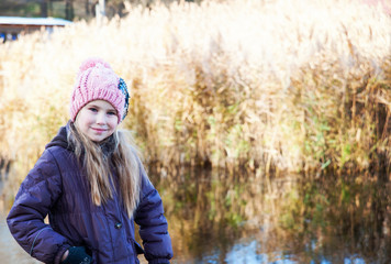 Fototapeta na wymiar Little beautiful blonde girl in pink hat on background of autumnal lake.