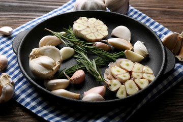 Fresh garlic with rosemary in black pan near blue checkered cotton napkin on wooden background, close up