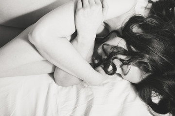 Lying on white bedsheet sensual curly brunette embracing her knees