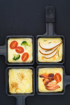 Four small square raclette pans with melted cheese and different ingredients: paprika, bacon, cherry tomatoes, broccoli and pear slices