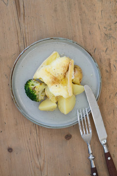 Plate with boiled potatoes and broccoli covered with melted raclette cheese on wooden background