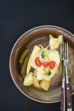 Plate with boiled potatoes and pickles covered with melted raclette cheese with cheery tomatoes and broccoli