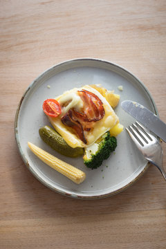 Plate with boiled potatoes, broccoli and pickles covered with melted raclette cheese with grilled bacon slice