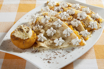 Portion of four pancakes stuffed with vanilla cream and peach, decorated with whipped cream and grated chocolate, served on a white plate.