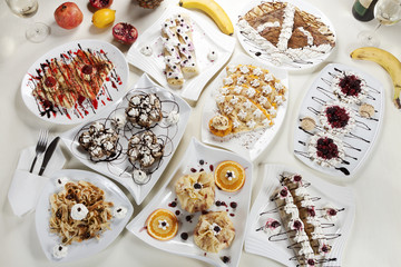 Different kinds of pancakes stuffed with various fillings and decorations, served on a table in white plates next to fresh fruit.