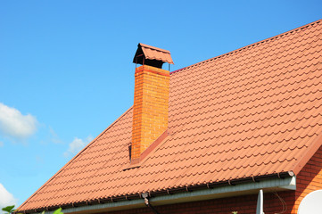 Faded red metal roof tile, rain gutter and chimney against blue sky. Bad roofing.