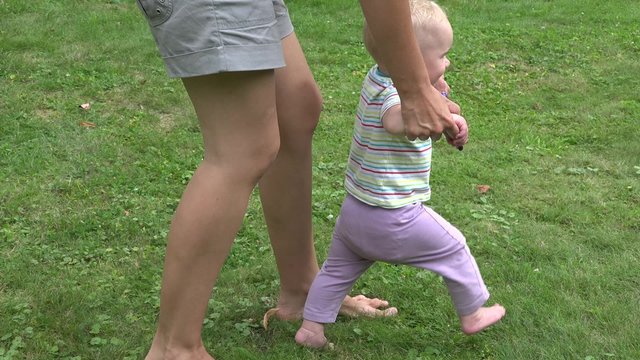 Cute funny happy baby making her first steps on green lawn in summer garden, mother holding his hands supporting by teaching to walk. Panorama shot. 4K