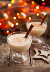 Eggnog traditional christmas egg, vanilla non alcohol liqueur preparation recipe in two glass cups with cinnamon sticks on wooden vintage table. Red bokeh background. shallow depth of field