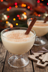 Eggnog traditional christmas egg, vanilla alcohol liqueur drink preparation recipe in two glass cups with cinnamon sticks on wooden vintage table. Red bokeh background. shallow depth of field