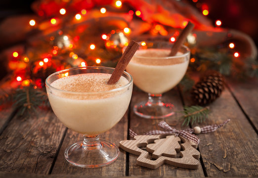 Eggnog Traditional Christmas Egg, Vanilla Rum Alcohol Drink Liqueur Preparation Recipe In Two Glass Cups With Cinnamon Sticks On Wooden Vintage Table. Red Bokeh Background. Shallow Depth Of Field