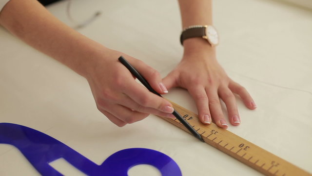 Fashion designer working on table. Hand of female tailor drawing pattern at paper in her studio. Young female tailor with measuring tape on shoulders working in workshop.