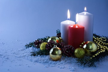 Glowing white and red Christmas candles, golden balls, fir branches and cones in snow on a light blue background