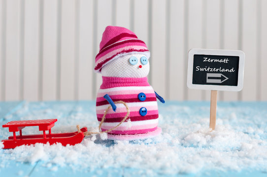 Signpost of the famous ski resort Zermatt, Switzerland and Snowman with red sled stand near direction sign. Postcard