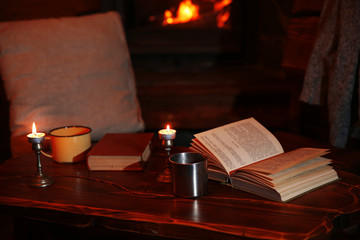 Fototapeta na wymiar Hot tea or coffee in mug, book and candles on vintage wood table. Fireplace as background