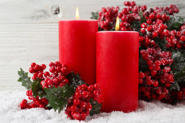 Candles and branch of holly berries in a snow over wooden background, close up