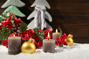 Fir trees, candles and balls with brunch of holly berries in a snow over wooden background, close up