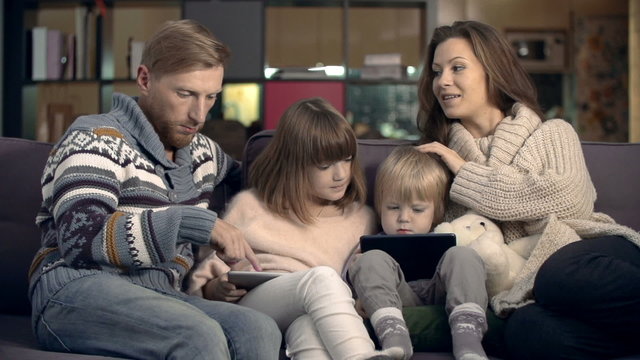 Happy family spending their evening together at home: kids playing on their tablets, mom and dad talking to each other