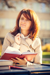  Campus.Portrait of a student thinking sitting on university bench outside studying with pen and book journal.Young business woman thinking writing diary at city park. Student.