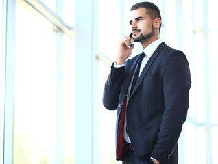 Portrait of a cheerful smiling men office worker talking on mobile phone while standing in modern office space, happy male entrepreneur having cell telephone conversation while resting after meeting