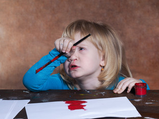 Маленький ребенок задумался. Девочка думает, что надо нарисовать. На столе бумага и краски. У девочки в руках кисть. Ребенок рисует красной краской. Девочка со светлыми волосами