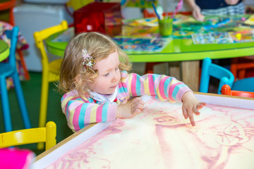 Fototapeta na wymiar Cute child girl drawing draws developing sand in preschool at table in kindergarten by method of Montessori