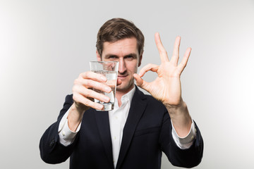 Blond-haired european businessman holding a glas of sparkling water with a satisfied look and giving a O.K. sign in front of a gradient background