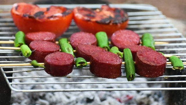 Turkish Sausage ( Sucuk ) cooking on the grill over coals. There are tomato and green pepper on the grill.
