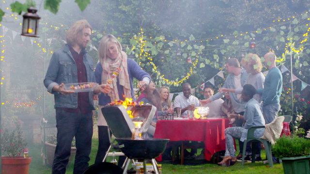 Happy attractive couple cooking at bbq while friends socialize in the background