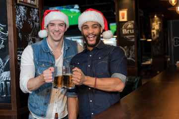 Two guys are standing with beer in a bar or pub wearing Santa Cl