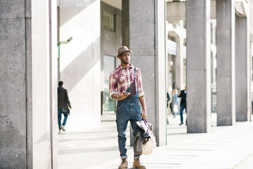 young handsome afro black man standing outside in the city, holding a bag and a smartphone, overlooking left - technology, social network, communication concept