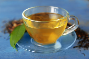 Glass cup of tea with green leaves and scattered tea around on blue wooden background