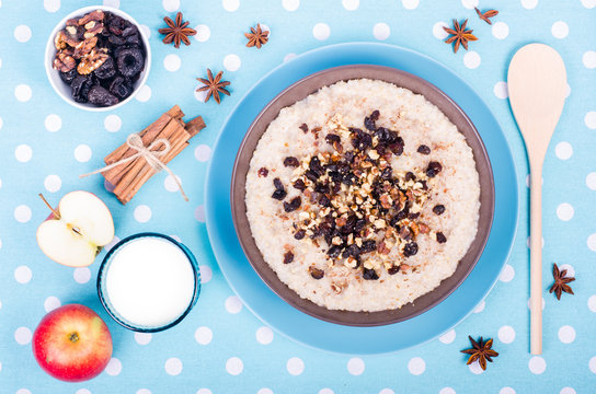 Breakfast set. A bowl of barley porridge with cocnut oil, vegetarian milk, walnuts, raisins, cinnamon on blue polka dot tablecloth, spoon, apple, star anise, glass of vegetarian milk.