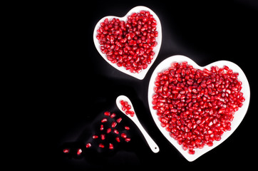 Two white heart shaped plates full of fresh ripe juicy pomegranate seeds, little spoon, whole fruit and ripe one on black background.