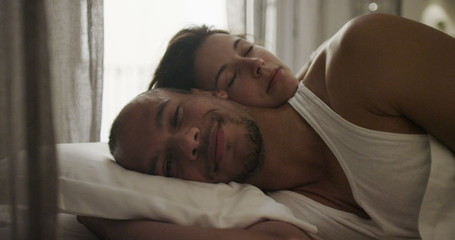 Happy sexy couple in the bedroom, man looking at camera while his partner sleeps