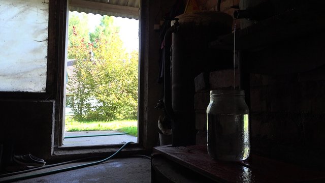 Traditional homemade distillery for making hooch vodka used in Europe countries. Alcohol drink flow in glass jar in rural farm room. Door to garden. Static closeup shot. 4K