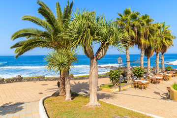 Fototapeta na wymiar Palm trees and restaurant tables on coastal promenade in Puerto de la Cruz town, Tenerife, Canary Islands, Spain