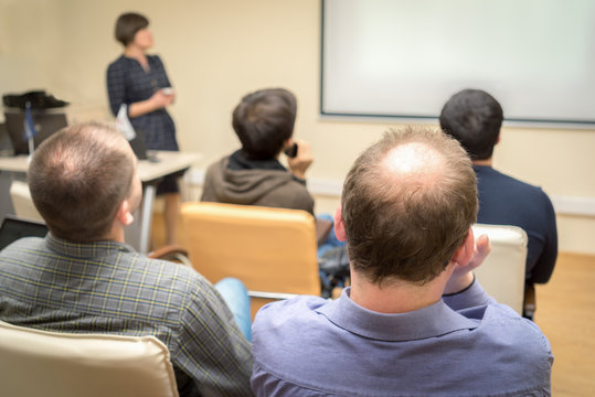 Back view of an adult students audience listening the presentation of a woman teacher near the screen