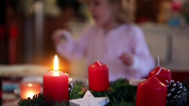 A close up of a burning candle on top of a wreath with a child nibble on some cookies in the background on advent first