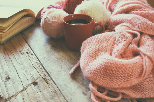 selective focus photo of pink cozy knitted scarf with to cup of coffee, wool yarn balls  and open book on a wooden table
