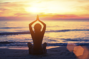 Silhouette of young girl in yoga pose sitting on the beach during sunset. - obrazy, fototapety, plakaty