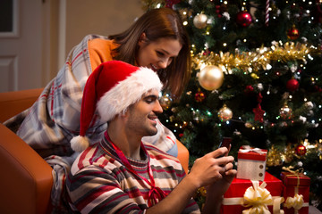 Young couple using smart phone while sitting in front of the Christmas tree