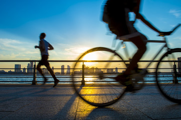 Silhouettes of jogger running and bicyclist cycling at sunset in front of the city skyline 