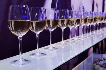 Nightclub wine glasses with white wine lit by party festive lights on dark-purple background,...