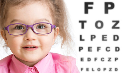 Smiling girl putting on glasses with blurry eye chart behind her - obrazy, fototapety, plakaty