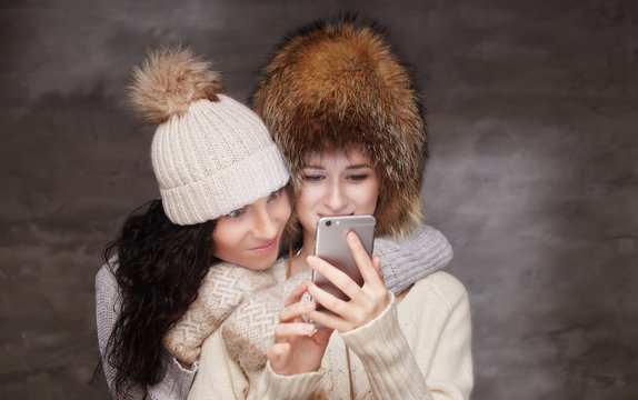 Two girls in fur winter hats looking at smartphone.
