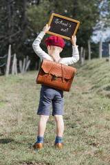 School Boy and his Chalkboard - School´s Out / Young school boy in vintage clothing and red beret hat with leather school bag on his back hold up a small blackboard over his head with text