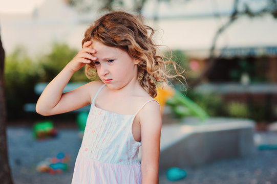 sad child girl with headache on summer playground, touching head