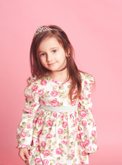 Smiling baby girl 4-5 year old posing in room over pink. Wearing trendy floral dress. Looking at camera.