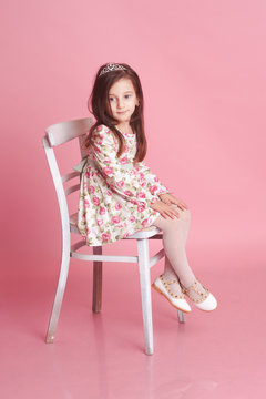 Cute baby girl 4-5 year old sitting on white chair over pink in room. Wearing floral pattern dress