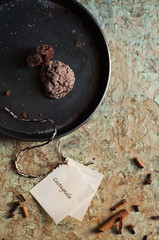 teglia nera con dolci speziati al cacao e caffè appena sfornati; fotografia zenitale con biscotto morsicato per mostrare l’interno, spezie sparse sulla base, etichetta con nastrino  